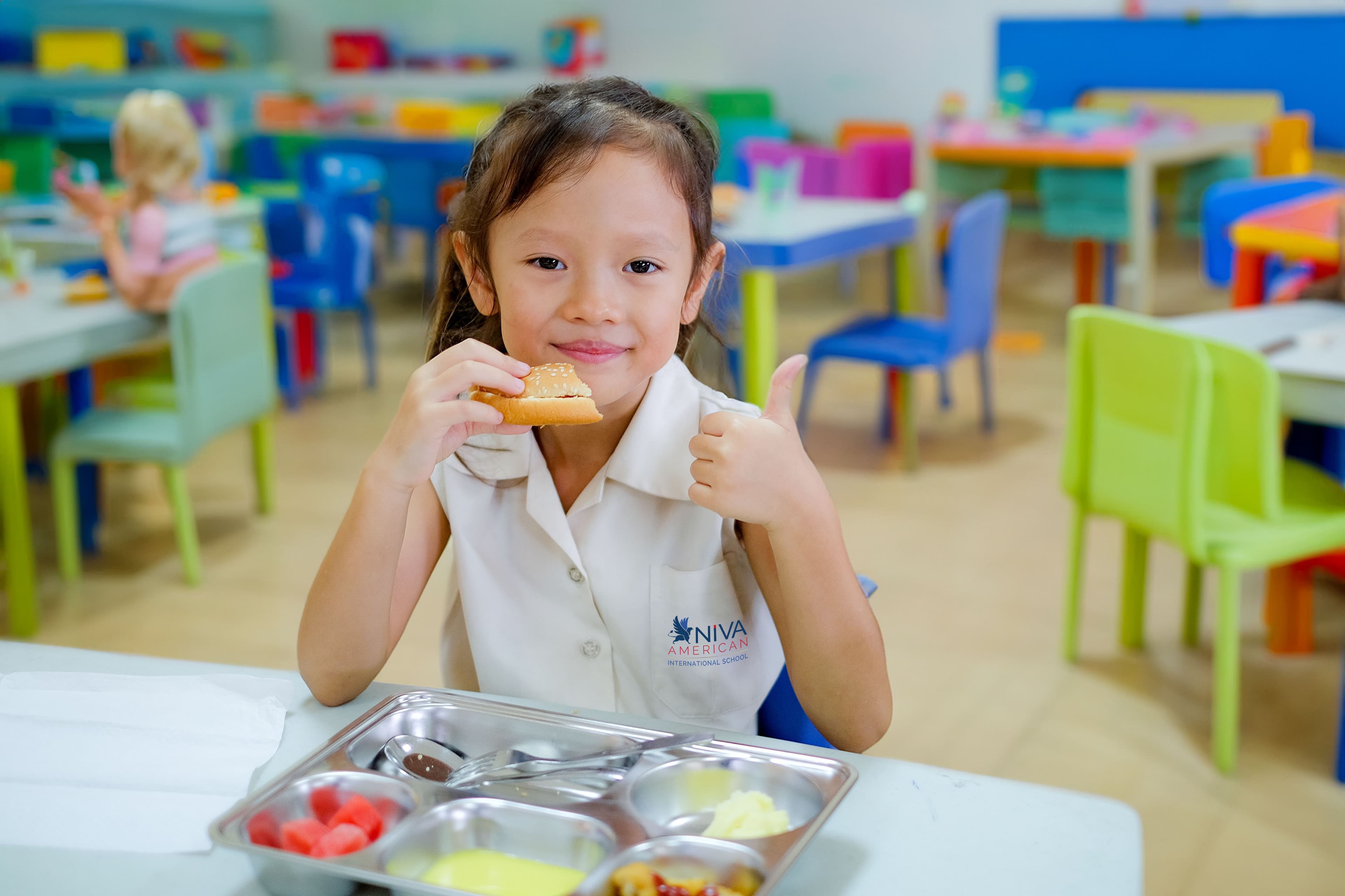 A NIVA American International School kindergarten student enjoying her lunch with a thumbs up